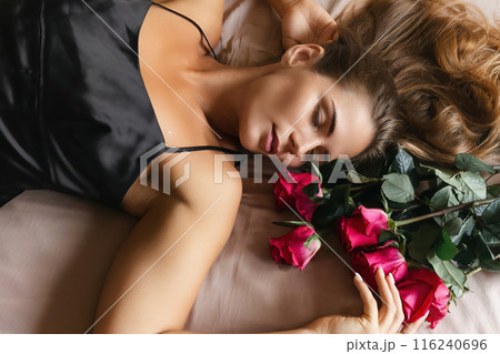 A woman is laying on a bed with a bouquet of red roses next to her. Concept of romance and intimacy, as the woman is surrounded by the flowers and he is in a relaxed and comfortable position. A woman is laying on a bed with a bouquet of red roses next to her. Concept of romance and intimacy, as the woman is surrounded by the flowers and he is in a relaxed and comfortable position. 116240696