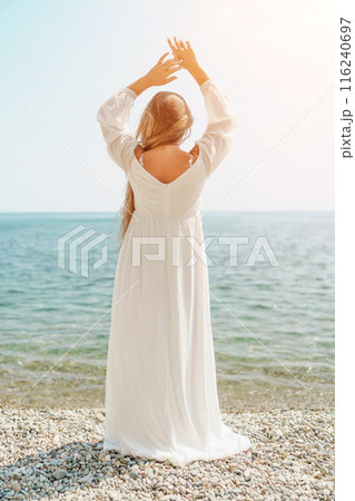 woman white dress stands on a beach, looking out at the ocean. She is in a contemplative or reflective mood, as she raises her hands above her head. Concept of peace and tranquility. 116240697