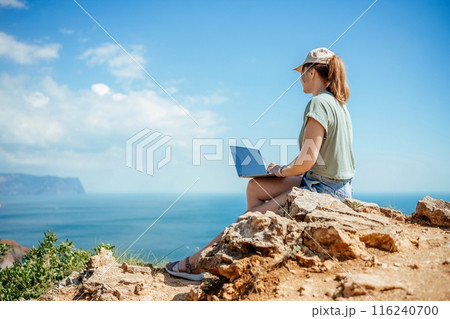 A woman is sitting on a rock overlooking the ocean with a laptop in front of her. She is working or studying, as she is focused on her laptop. Concept of solitude and tranquility. A woman is sitting on a rock overlooking the ocean with a laptop in front of her. She is working or studying, as she is focused on her laptop. Concept of solitude and tranquility. 116240700