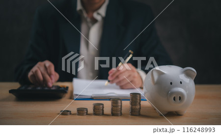 Businessman calculates finances with a piggy bank and stacks of coins, saving money wealth and financial planning. Business investment and tax concept Businessman calculates finances with a piggy bank and stacks of coins, saving money wealth and financial planning. Business investment and tax concept 116248993