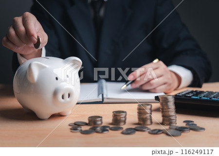 Person in suit depositing coin into piggy bank on desk with coins and calculator. investment and save money tax finance management concept 116249117