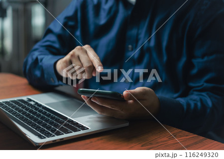 Close-up of a businessperson multitasking with a mobile phone and laptop at a wooden desk Close-up of a businessperson multitasking with a mobile phone and laptop at a wooden desk 116249320