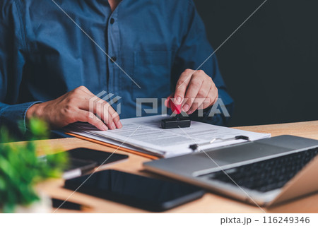 A person's hands are seen stamping a document, symbolizing the approval process in a professional setting. The laptop in the background suggests a modern office environment. A person's hands are seen stamping a document, symbolizing the approval process in a professional setting. The laptop in the background suggests a modern office environment. 116249346