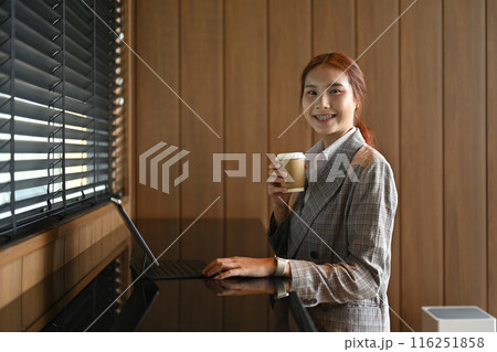 Young businesswoman holding paper cup and using digital tablet in modern office 116251858
