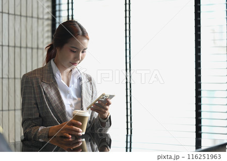 Portrait of Asian businesswoman in formalwear holding paper cup and texting on a mobile app 116251963