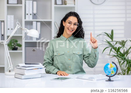 Cheerful female teacher raising her hand while sitting at a desk in a modern, organized classroom. Surrounded by books, a globe, and office supplies, she exudes enthusiasm and knowledge ready teach. Cheerful female teacher raising her hand while sitting at a desk in a modern, organized classroom. Surrounded by books, a globe, and office supplies, she exudes enthusiasm and knowledge ready teach. 116256566