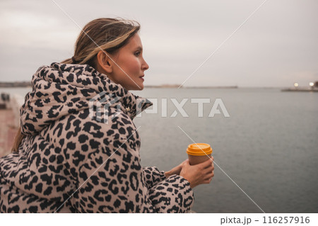 A woman in a leopard print coat is holding a coffee cup and looking out at the water. A woman in a leopard print coat is holding a coffee cup and looking out at the water. 116257916