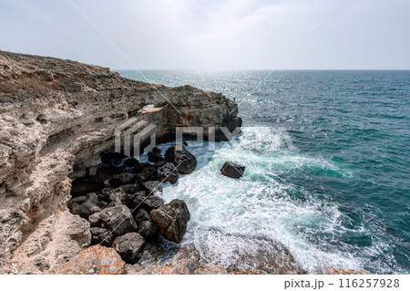The ocean is calm and the sky is cloudy. The water is a deep blue color. The rocks are jagged and the waves are small. 116257928