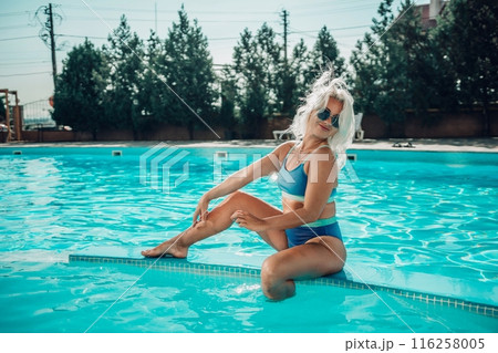 A blonde woman is sitting on a pool ledge in a blue bikini. She is smiling and looking at the camera. 116258005