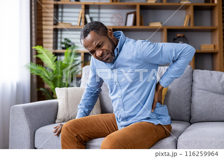 A man is sitting on a couch in a modern home interior, holding his back and expressing discomfort. The image captures the concept of back pain, discomfort, and health issues within a home setting. 116259964
