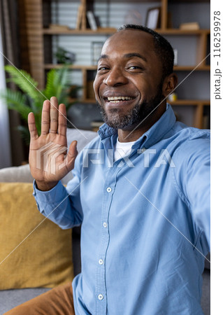 A cheerful man wearing a blue shirt waves and smiles during a video call from his living room. The warm and cozy environment suggests a relaxed and friendly conversation. A cheerful man wearing a blue shirt waves and smiles during a video call from his living room. The warm and cozy environment suggests a relaxed and friendly conversation. 116259978