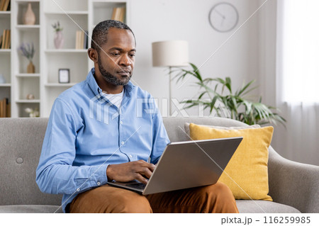A man working from home on his laptop while sitting on a comfortable sofa in a well-decorated living room. The setting is relaxed, promoting productivity and focus. 116259985