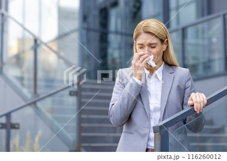 A professional woman in a business suit is sneezing while holding a tissue outside a contemporary office building. She appears to be experiencing allergy symptoms or a cold. 116260012