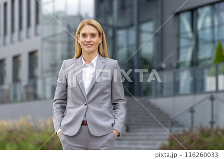 Portrait of a confident businesswoman in a formal suit standing outdoors in front of a modern office building. The woman is smiling, exuding professionalism and success in a corporate environment. 116260033