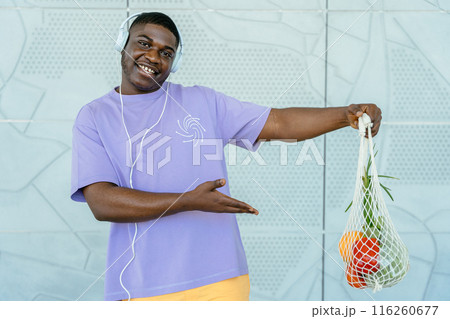 Portrait of happy young black man showing mesh bag of fresh groceries he bought at local market. Eco friendly (zero waste) lifestyle and healthy diet concept. 116260677