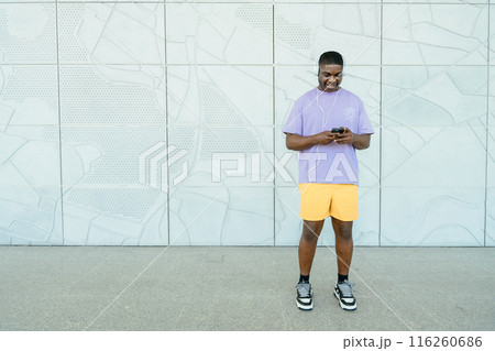 Cheerful young man in sportwear using smartphone while stands against white wall urban background. Full height portrait 116260686