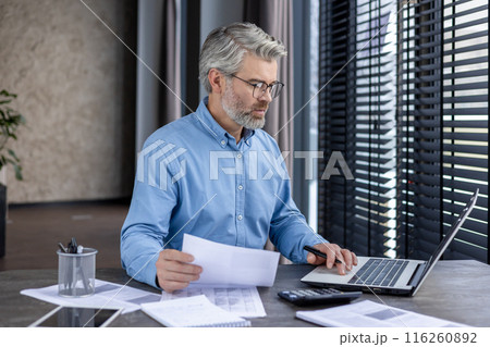 Mature businessman wearing glasses, working on laptop at a desk with documents and calculator in a modern office. Focused professional analyzing data, financial reports, and doing paperwork. 116260892