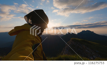 A man in a yellow windbreaker and head baffle stands against a mountainous landscape and watches the sunset. The serene beauty of nature is captured in this serene evening scene. A man in a yellow windbreaker and head baffle stands against a mountainous landscape and watches the sunset. The serene beauty of nature is captured in this serene evening scene. 116275753