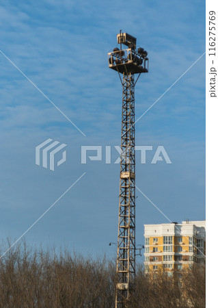 A tall metal lattice tower with floodlights against a blue sky. The tower is constructed of metal crossbeams. Cityscape, a multi-storey apartment block is seen on the right. 116275769