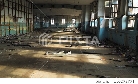 The interior of an abandoned dilapidated building with broken windows, peeling paint and rubbish scattered on the floor. Sunlight streams through the windows, casting shadows across the room. 116275771