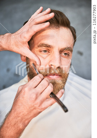 Isolated photo of handsome man getting beard cut. Professional hairdresser using beard blade for shaping. Isolated photo of handsome man getting beard cut. Professional hairdresser using beard blade for shaping. 116277903