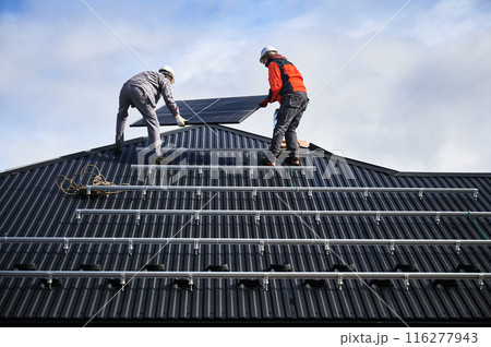 Electricians installing solar panel system on roof of house. Men workers in helmets carrying photovoltaic solar module outdoors. Concept of alternative and renewable energy. 116277943