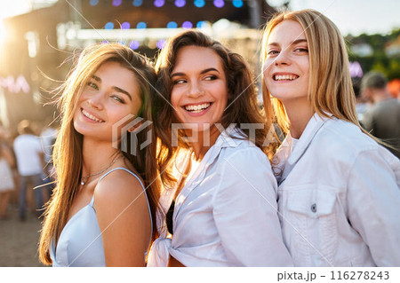 Three smiling young women enjoy summer music festival on sandy shore. Friends pose in sunlight, stage lights behind. Casual fashion, fun, togetherness at outdoor concert event by sea. Three smiling young women enjoy summer music festival on sandy shore. Friends pose in sunlight, stage lights behind. Casual fashion, fun, togetherness at outdoor concert event by sea. 116278243