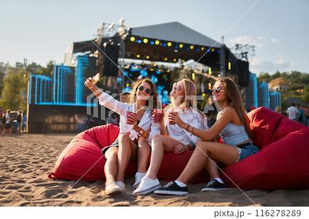 Three women sit on red bean bags, sunlit beach as backdrop, holding drinks, enjoying music festival. Smiling ladies in sunglasses relax by stage, casual summer outfits, friends at seaside. 116278289