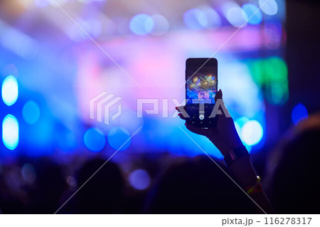 Concertgoer films live music event with smartphone, hand raised above crowd, capturing stage lights, performers in blurred background. Energetic festival atmosphere, fans enjoy night performance. Concertgoer films live music event with smartphone, hand raised above crowd, capturing stage lights, performers in blurred background. Energetic festival atmosphere, fans enjoy night performance. 116278317