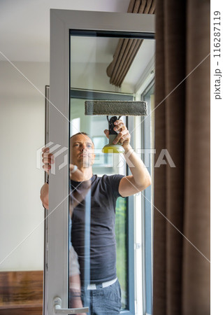 Young man cleans the windows in the house. He splashes detergent, rubs and removes water at the glass Young man cleans the windows in the house. He splashes detergent, rubs and removes water at the glass 116287119