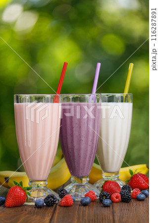 set of milkshakes in glasses with straw and fresh bananas and berries on table outdoors set of milkshakes in glasses with straw and fresh bananas and berries on table outdoors 116287891