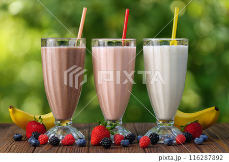set of milkshakes in glasses with straw and fresh bananas and berries on table outdoors 116287892