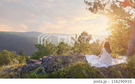 Woman in white dress meditating on a rocky cliff during sunset with forested hills in the background. Woman in white dress meditating on a rocky cliff during sunset with forested hills in the background. 116290910