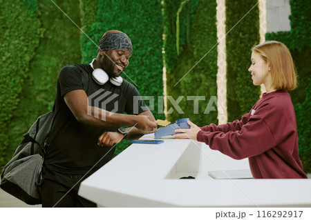 Buff African American man making payment for VIP membership card and tapping his golden card while female receptionist holding payment terminal 116292917