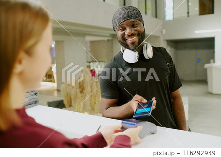 Young African American man paying for his new gym subscription via his phone, he smiling widely to female manager Young African American man paying for his new gym subscription via his phone, he smiling widely to female manager 116292939