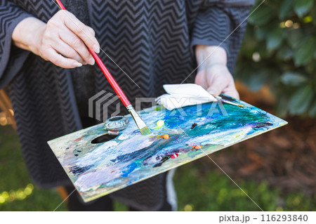 Close up of the palette and hands of an elderly woman artist painting a picture in outdoors. Close up of the palette and hands of an elderly woman artist painting a picture in outdoors. 116293840