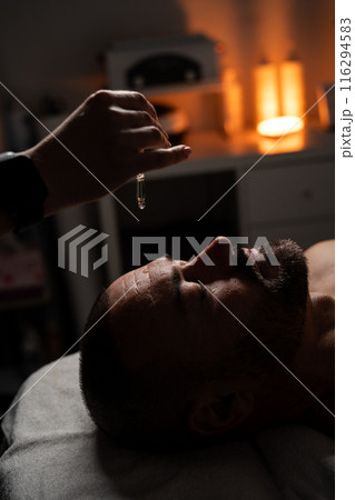 Oil is dripped onto a man's forehead for massage. Close up of a handsome man enjoying facial massage at spa center. Oil is dripped onto a man's forehead for massage. Close up of a handsome man enjoying facial massage at spa center. 116294583