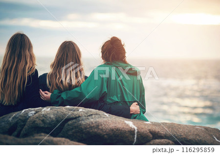 Friends, beach and sunset of people sitting on rock for bonding, back view and embrace in Ireland. Vacation, group and relax for fun day by ocean, nature or contemplation in outdoors for togetherness 116295859