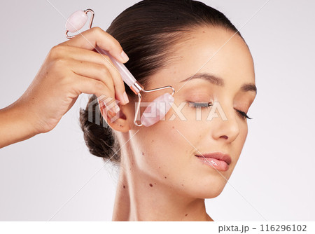 Skincare, roller and woman in studio with eyes closed for facial treatment, massage on white background. Stone, gua sha and hand of model with tool for rose quartz, dermatology or lymph cleaning 116296102