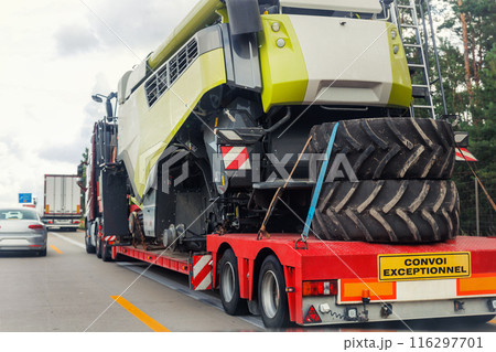 Long heavy industrial truck with semi trailer platform transport disassembled combine harvester machine on common highway on bright summer day. Agricultural equipment transportation service Long heavy industrial truck with semi trailer platform transport disassembled combine harvester machine on common highway on bright summer day. Agricultural equipment transportation service 116297701