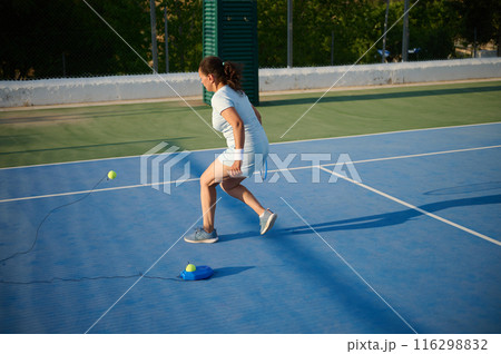 Woman practicing tennis on court with ball trainer under sunlight Woman practicing tennis on court with ball trainer under sunlight 116298832