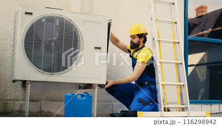 Professional worker starting work on malfunctioning air conditioner, using power drill to take apart condenser side coil panel. Capable electrician dismantling hvac system to check for faulty wires 116298942