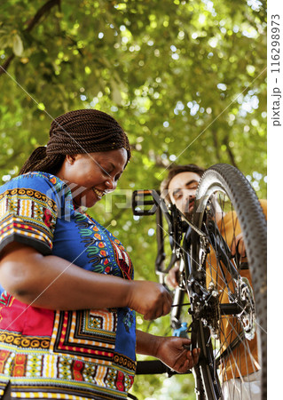 Sport-loving enthusiastic black woman fixing bike derailleur in yard for summer leisure cycling. Healthy and active african american female cyclist receiving help from caucasian man for bicycle 116298973