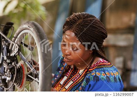 Dedicated sports-loving black woman meticulously inspects her bike chain using professional tool. Young african american lady adjusting and securing modern bicycle parts as summer maintenance activity Dedicated sports-loving black woman meticulously inspects her bike chain using professional tool. Young african american lady adjusting and securing modern bicycle parts as summer maintenance activity 116299007