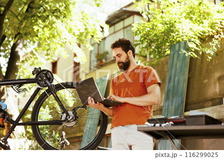 Active male caucasian cyclist using laptop to search for online repair guides while mending his broken bicycle chain. Sports-loving man upkeeping and servicing bike by browsing the internet on Active male caucasian cyclist using laptop to search for online repair guides while mending his broken bicycle chain. Sports-loving man upkeeping and servicing bike by browsing the internet on 116299025