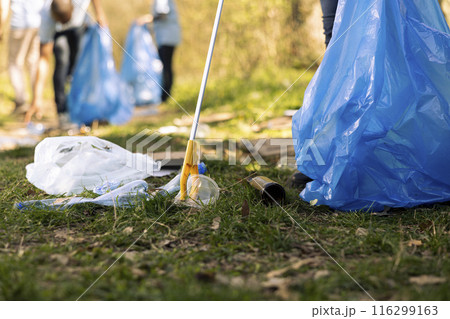 Activist using claw tool to collect rubbish and plastic junk from the ground, storing garbage in a trash bag and clean the natural forest ecosystem. Male environmentalist gathering litter. 116299163