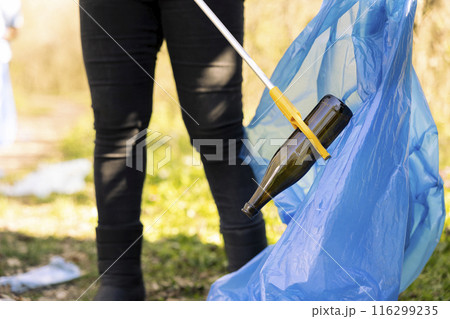 Woman activist grabbing rubbish with a claw tool in the forest, clearing ecosystem of trash and plastic junk. Female activist doing voluntary work to combat illegal dumping, save the planet. Woman activist grabbing rubbish with a claw tool in the forest, clearing ecosystem of trash and plastic junk. Female activist doing voluntary work to combat illegal dumping, save the planet. 116299235