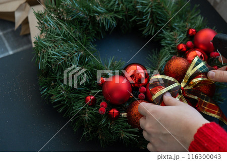Woman tying decorative ribbon Step by step instruction of making handmade Christmas wreath made of festive decorations in red color. Female hands creating craft homemade decor. New year celebration 116300043