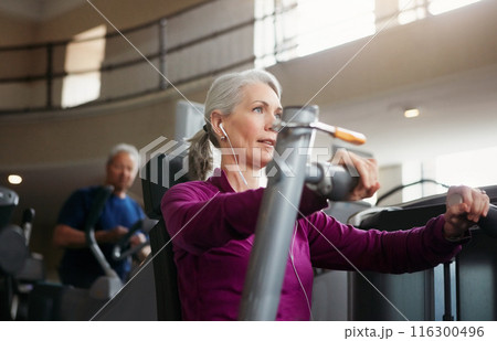 Mature woman, chest press and music with machine for fitness, workout or indoor exercise at gym. Active female person with earphones on equipment for strength, muscle or wellness at health club 116300496