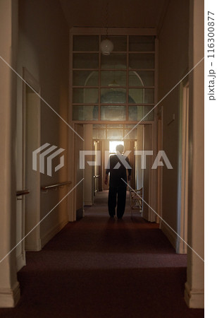 Back, stick and old woman in home walking in living room in retirement in dark hallway or house. Healthy, senior lady and elderly female person with disability, calm peace or cane to relax or balance 116300877